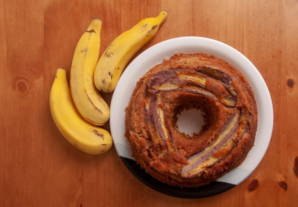 bolo de banana sobre um prato branco com três bananas ao redor da mesa