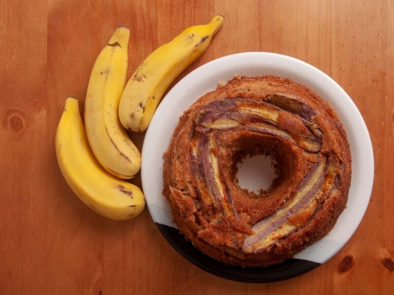 bolo de banana sobre um prato branco com três bananas ao redor da mesa